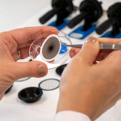 a closeup image of hands holding tweezers and a clear dish containing a paper-like disk with a black mark in its centre 